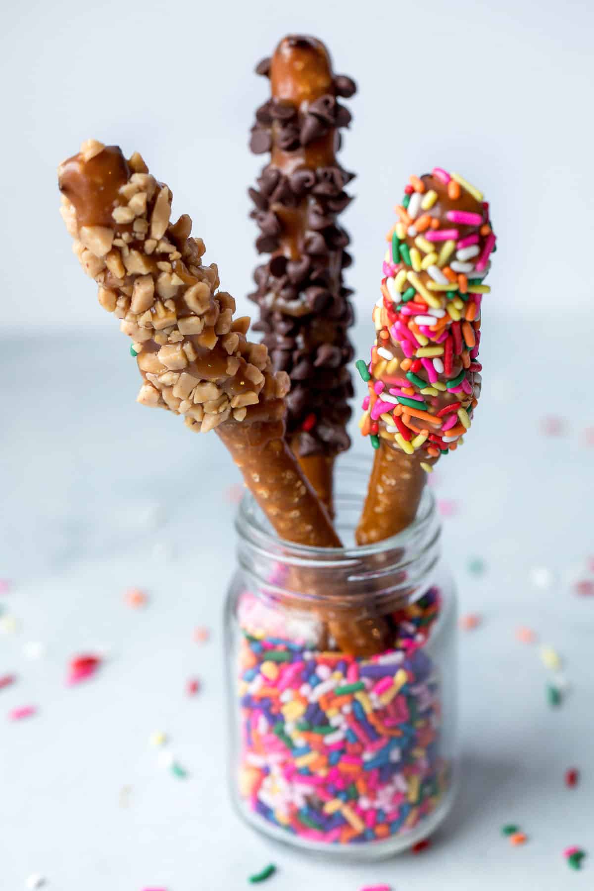 3 pretzel rods dipped in caramel and various toppings standing up in a glass jar with sprinkles