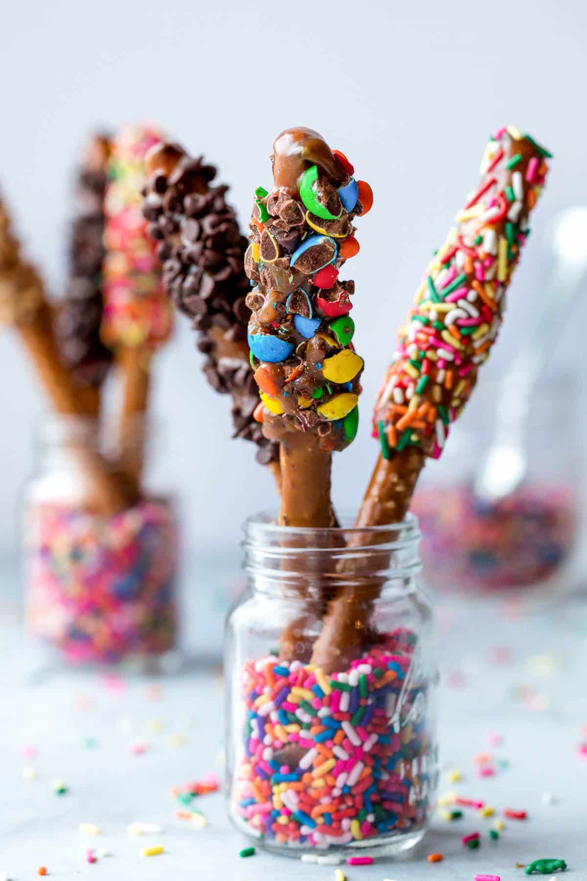 pretzel rods dipped in caramel and various toppings standing up in glass jars with sprinkles