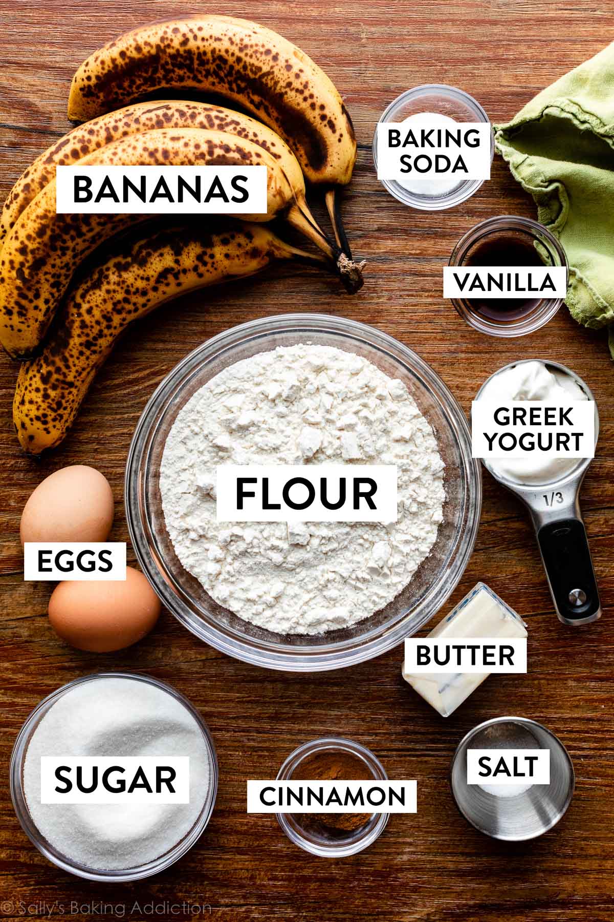 flour, butter, Greek yogurt, vanilla, baking soda, bananas, eggs, and other ingredients on wooden backdrop.