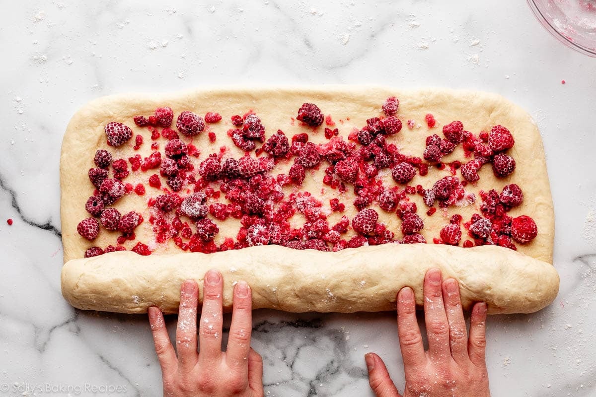 hands rolling up dough with frozen raspberries on top.
