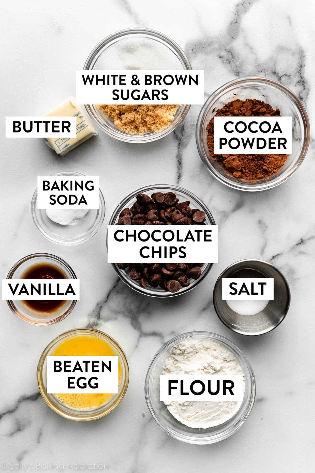 bowls of ingredients on marble counter including cocoa powder, salt, flour, chocolate chips, beaten egg, sugars, and more.