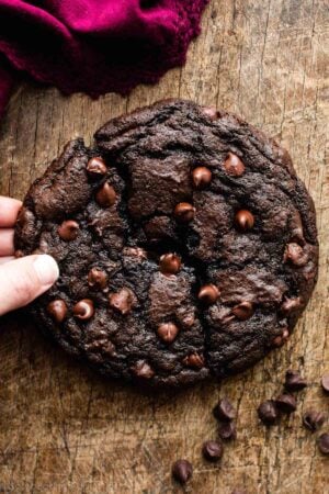 one giant double chocolate cookie on wooden cutting board with hand pulling broken half away.