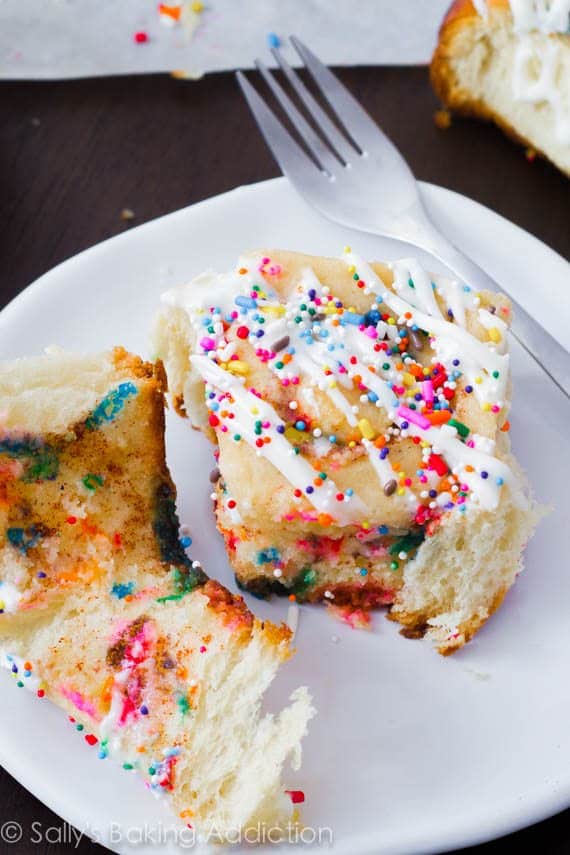 cake batter cinnamon rolls topped with vanilla glaze and sprinkles on a white plate with a fork