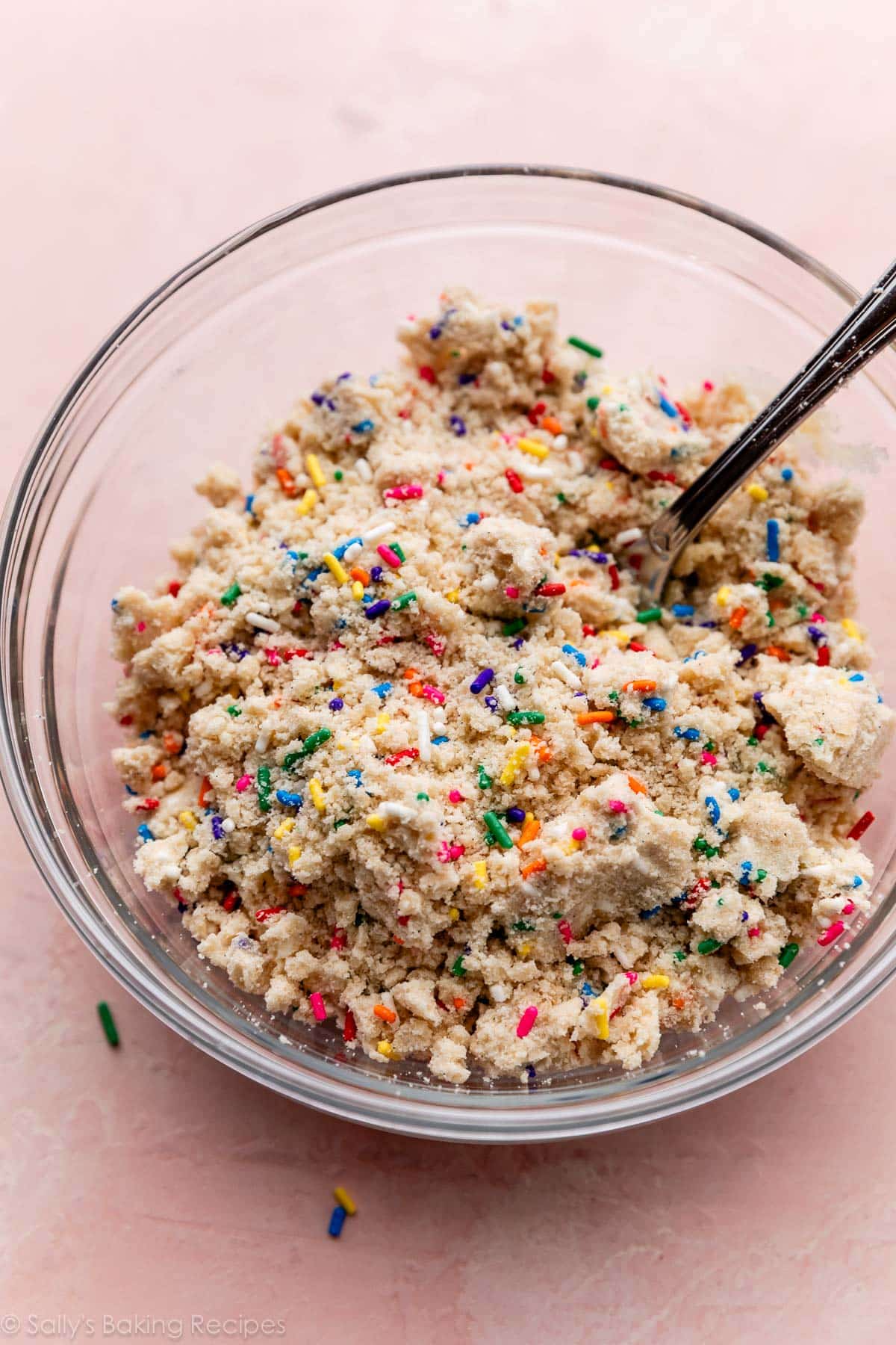 crumble mixture with rainbow sprinkles in glass bowl on pink surface.