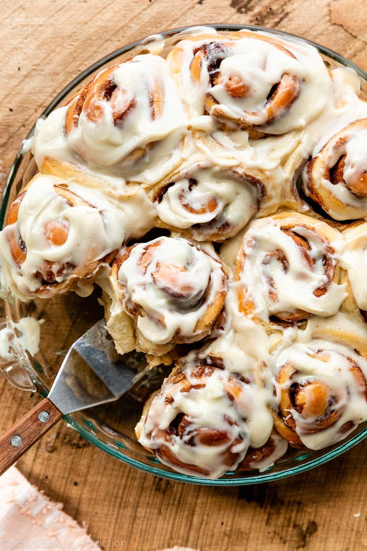 round pan of cinnamon rolls with cream cheese icing and a spatula removing one.
