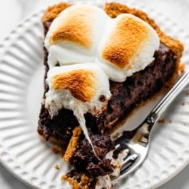 forkful and slice of s'mores brownie pie with toasted marshmallows on top on white plate.
