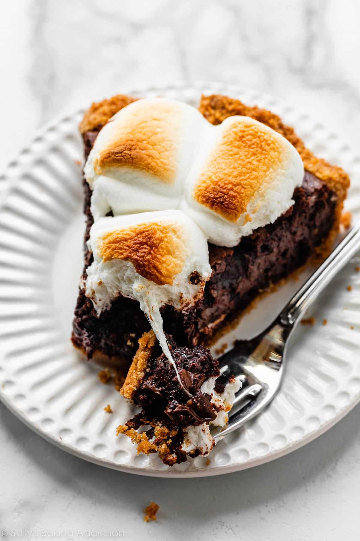 forkful and slice of s'mores brownie pie with toasted marshmallows on top on white plate.