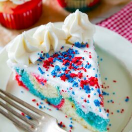 slice of red, white, and blue tie-dye cake topped with vanilla frosting and sprinkles on a white plate with a fork