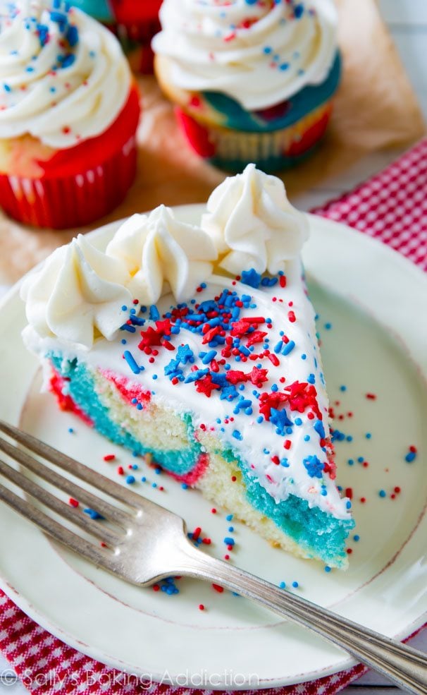 slice of red, white, and blue tie-dye cake topped with vanilla frosting and sprinkles on a white plate with a fork