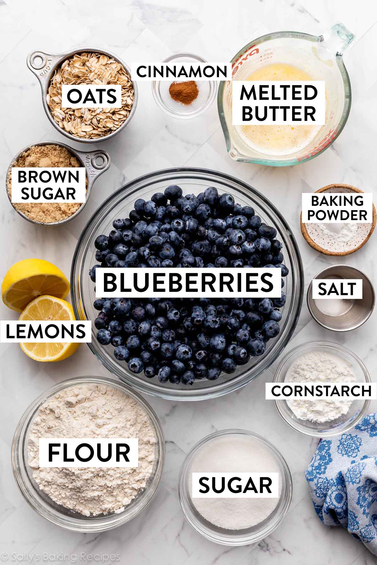ingredients in separate bowls on marble counter including blueberries, sugar, flour, lemon, melted butter, oats, brown sugar, and more.