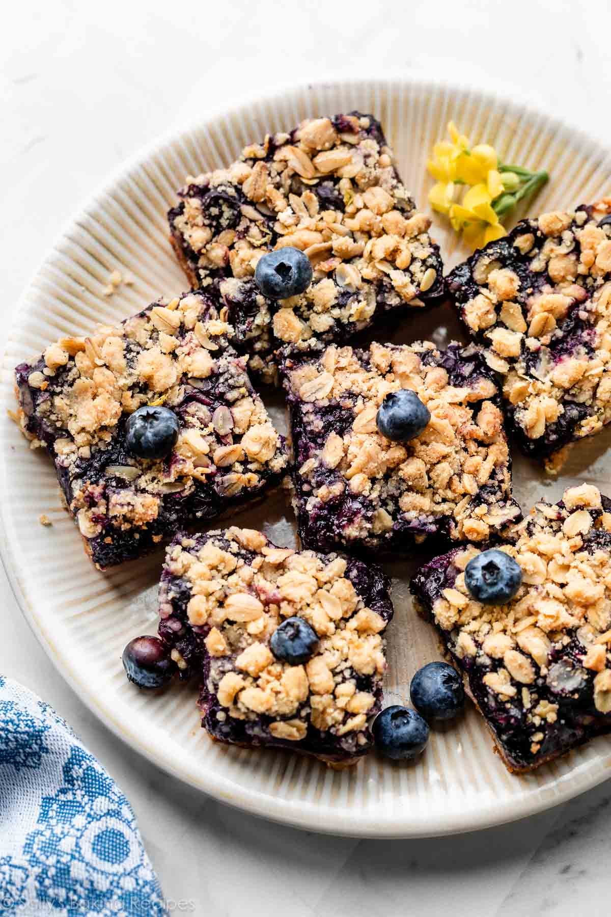 blueberry pie bars arranged on beige plate with fresh blueberries dotted on top.