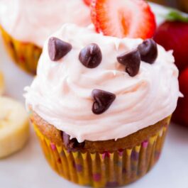 banana chocolate chip cupcakes topped with strawberry cream cheese frosting, chocolate chips, and strawberry slices on a white plate