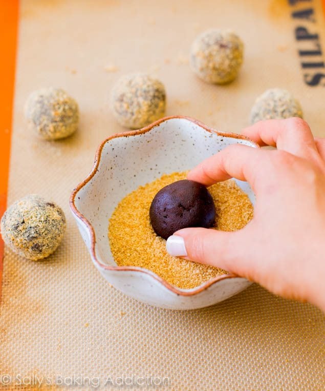 hand rolling chocolate crinkle cookie dough ball in a bowl of graham cracker crumbs in a bowl