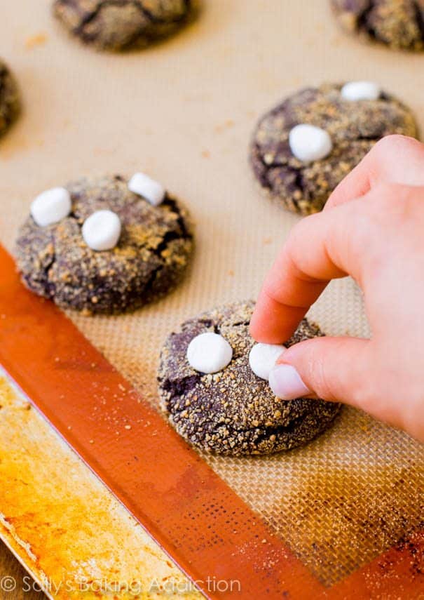 hand placing marshmallows on top of s'mores chocolate crinkle cookies on a silpat baking mat