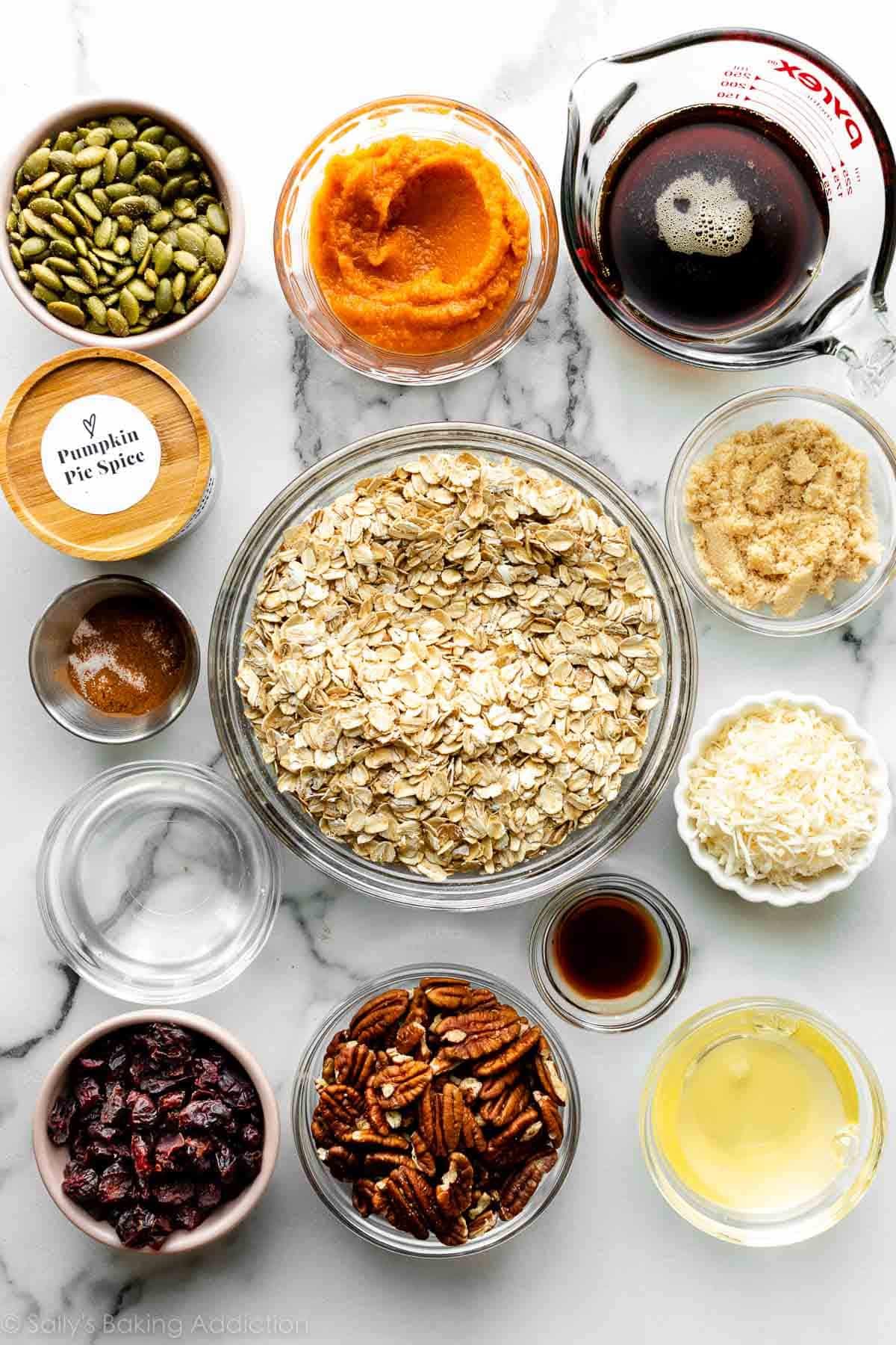 many measured ingredients on marble counter including maple syrup, pumpin, oats, pepitas, egg whites, pecans, coconut, and more.