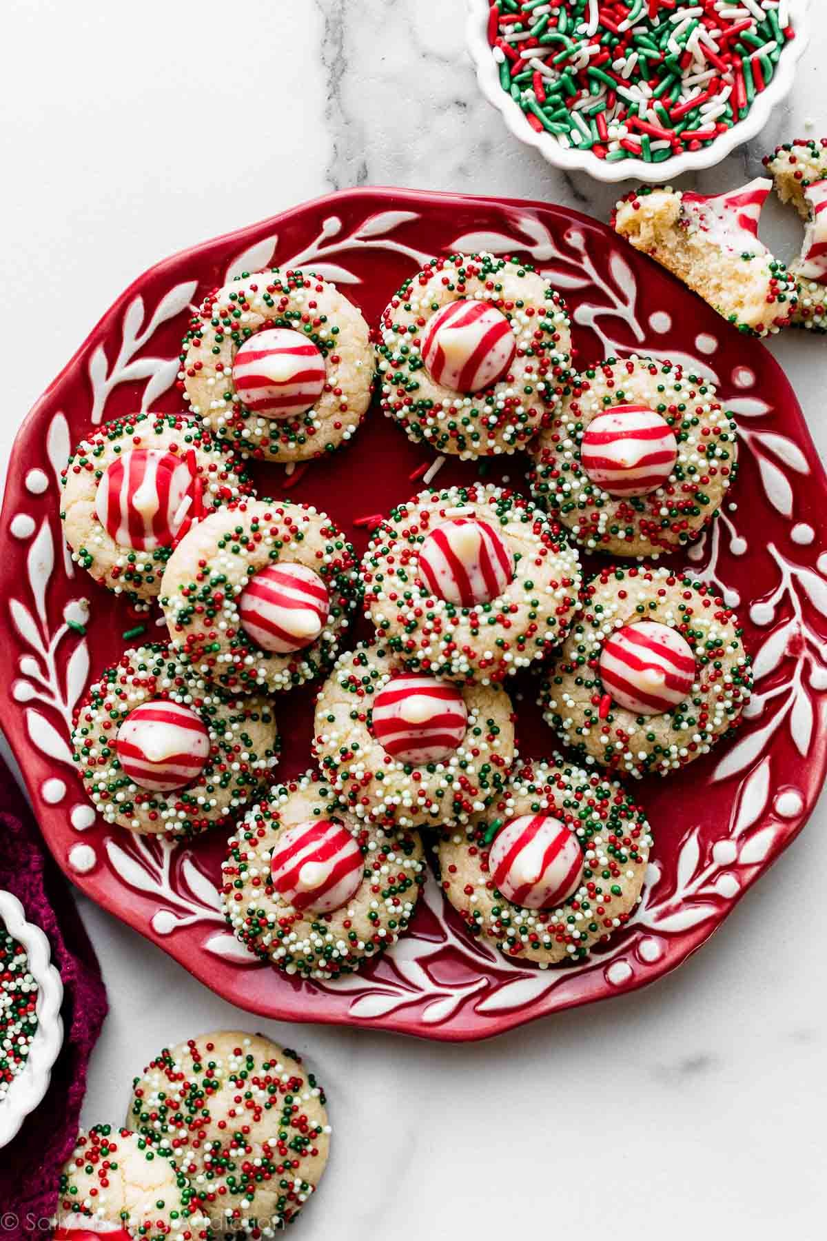 11 sugar cookies rolled in Christmas nonpareil sprinkles with candy cane Hershey's Kiss pressed into center arranged on red plate.