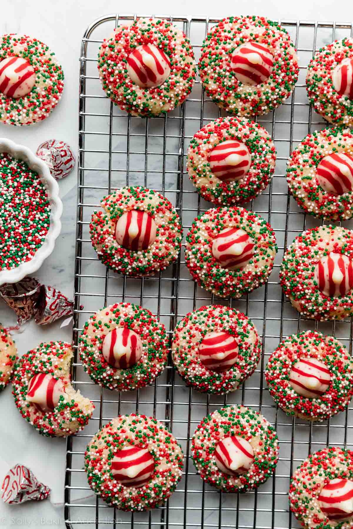 sprinkle-covered sugar cookies with candy cane Hershey's Kiss in the center of each arranged on cooling rack.