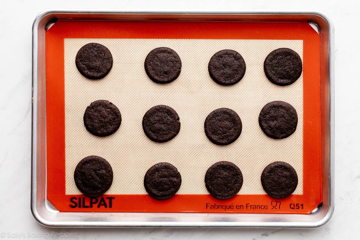 12 flattened chocolate cookies on Silpat-lined baking sheet.