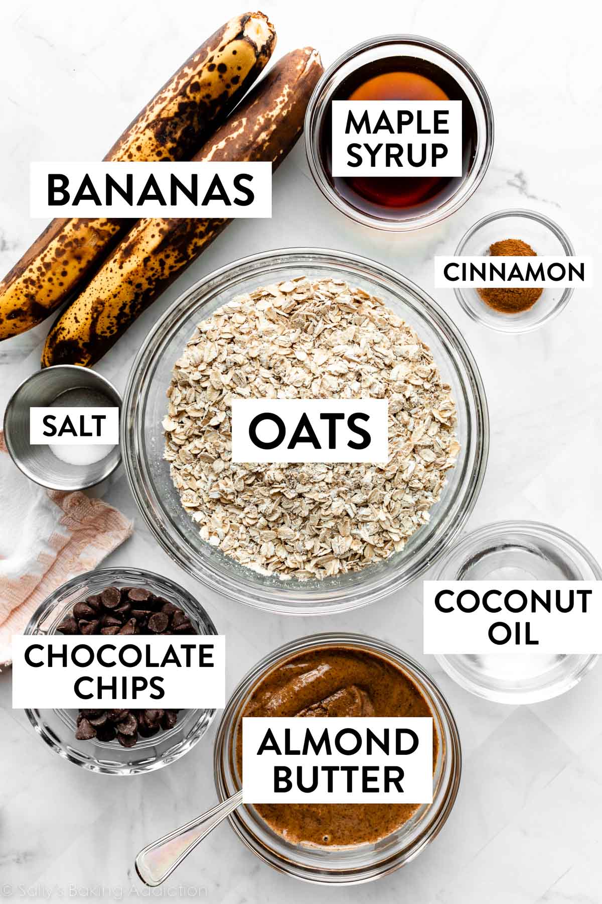 ingredients measured on counter including maple syrup, cinnamon, coconut oil, almond butter, 2 bananas, chocolate chips, and oats.