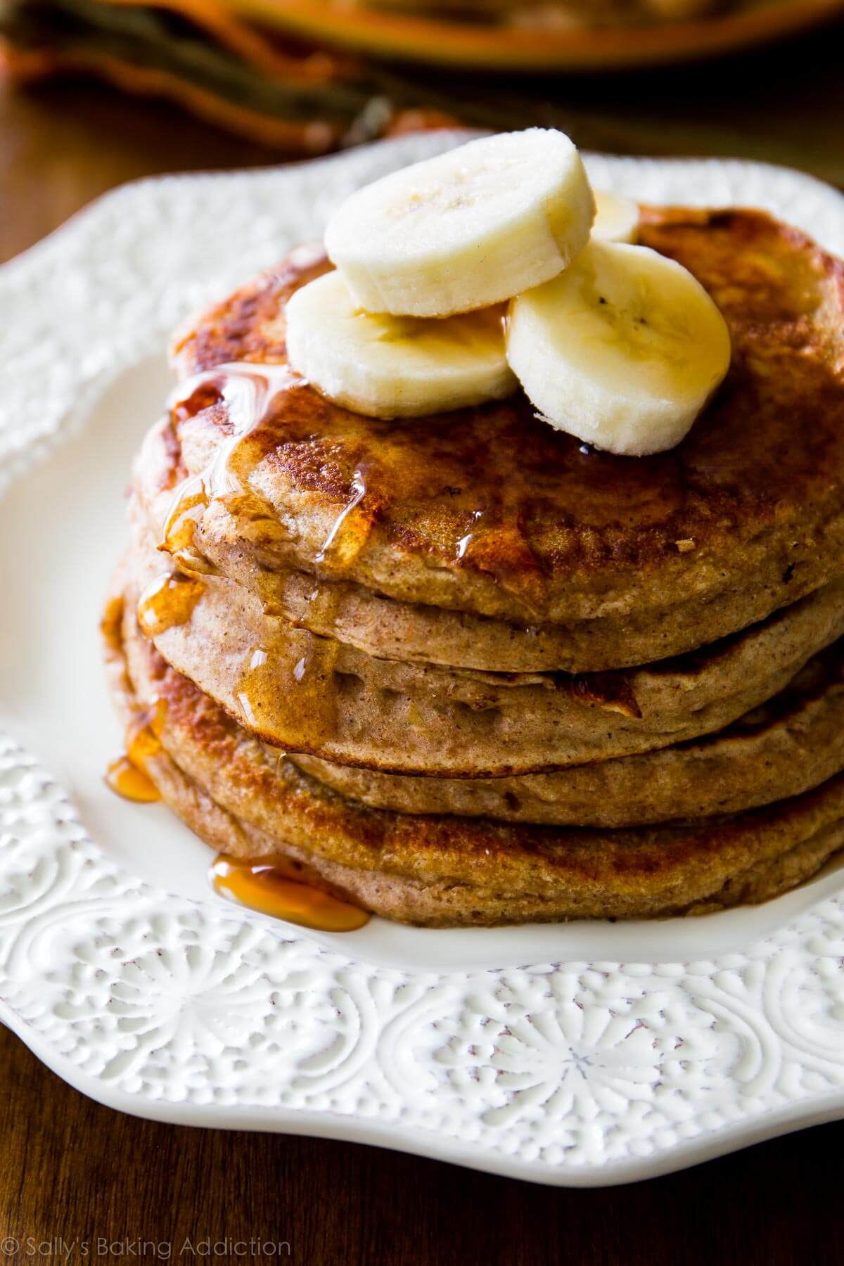 stack of whole wheat banana pancakes on a white plate topped with maple syrup and banana slices