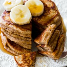 stack of whole wheat banana pancakes on a white plate topped with maple syrup and banana slices