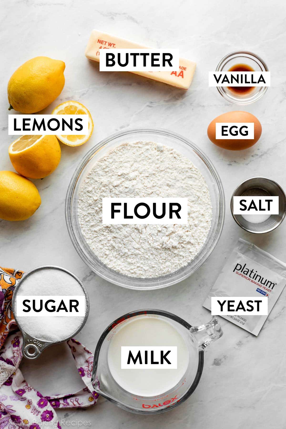 ingredients on marble counter including flour, salt, egg, vanilla, butter, lemons, sugar, and milk.