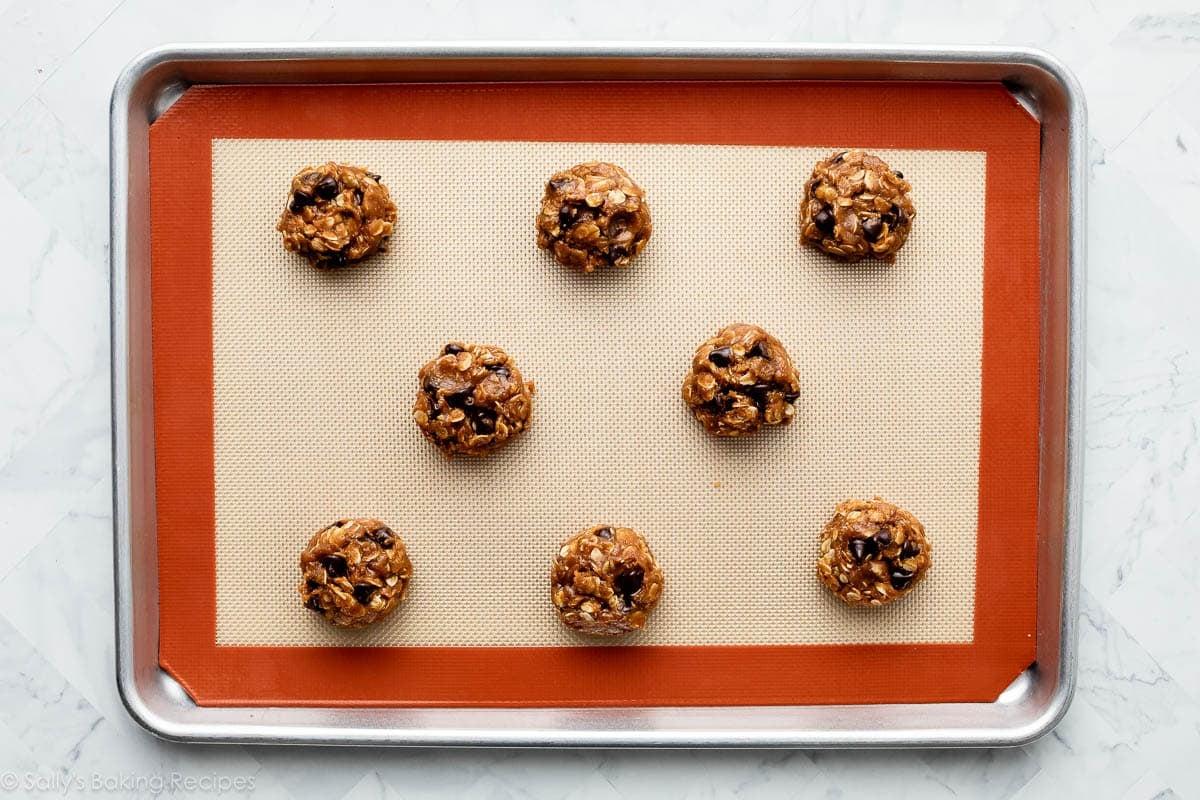 8 oatmeal cookie dough balls arranged on silicone baking mat-lined baking sheet.