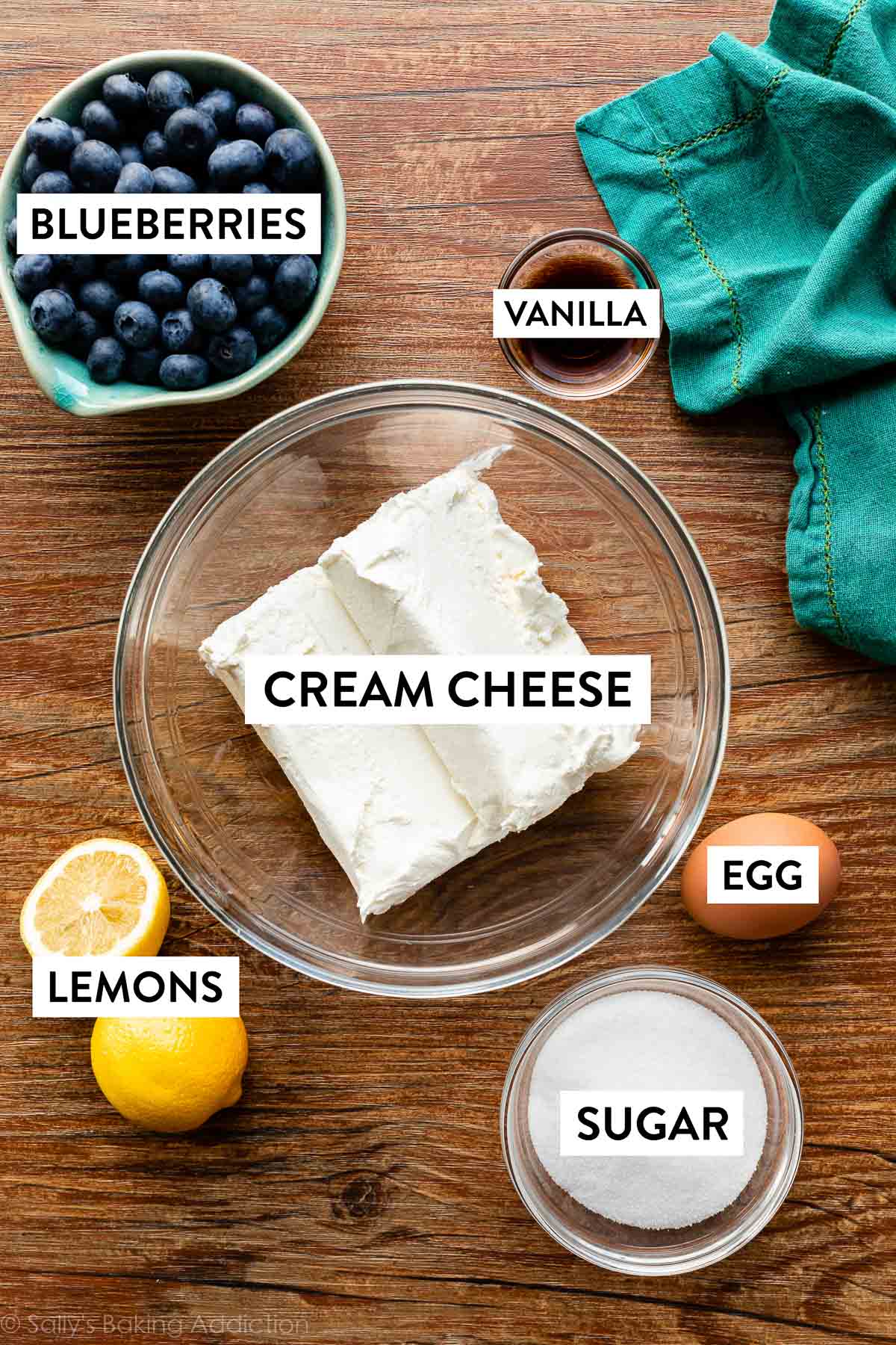 ingredientes en tablero de madera incluyendo queso crema, azúcar, huevo, vainilla, arándanos y limón.