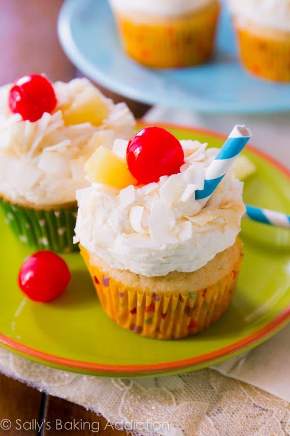 piña colada cupcakes topped with coconut frosting, shredded coconut, a cherry, slice of pineapple, and a straw on a green plate