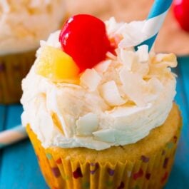 piña colada cupcakes topped with coconut frosting, shredded coconut, a cherry, slice of pineapple, and a straw