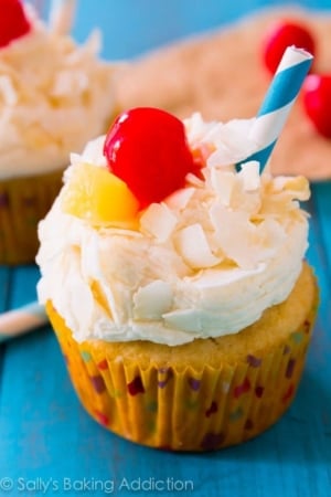 piña colada cupcakes topped with coconut frosting, shredded coconut, a cherry, slice of pineapple, and a straw