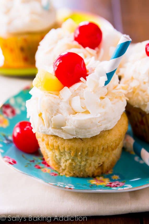 piña colada cupcakes topped with coconut frosting, shredded coconut, a cherry, slice of pineapple, and a straw on a floral plate