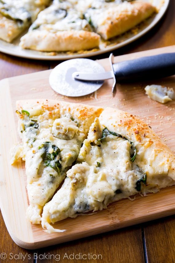 slices of spinach artichoke pizza on a wood cutting board with a pizza cutter