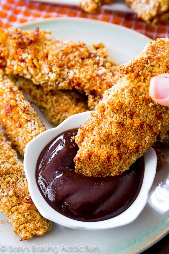hand dipping baked chicken finger into bbq sauce in a white bowl