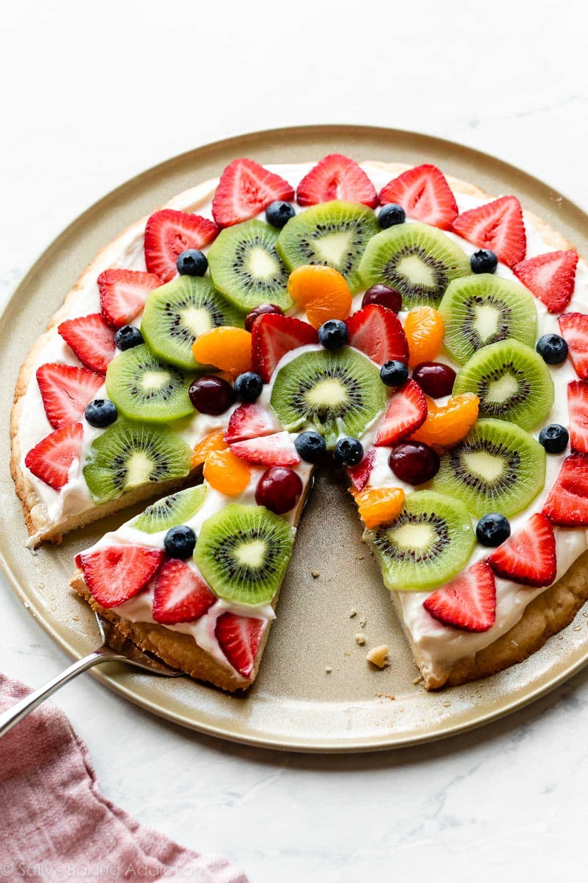 decorated sugar cookie fruit pizza on pizza pan with strawberries, kiwi, blueberries, grapes, and mandarin oranges on top.