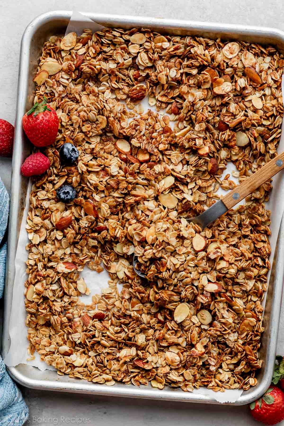 homemade vanilla almond granola spread on lined baking sheet with a couple fresh strawberries and blueberries.