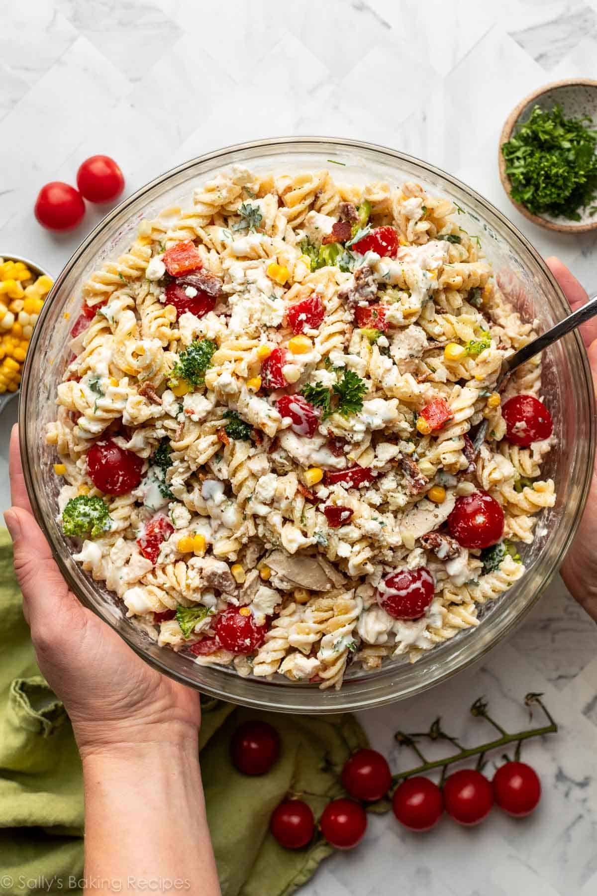 hands picking up glass bowl of pasta salad with tomatoes, broccoli, corn, and red bell peppers.
