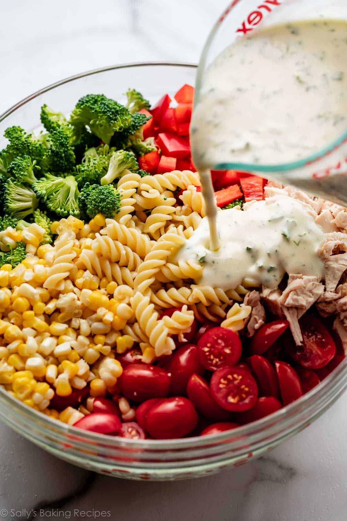 pouring creamy dressing on bowl of pasta, tomatoes, corn, and broccoli.