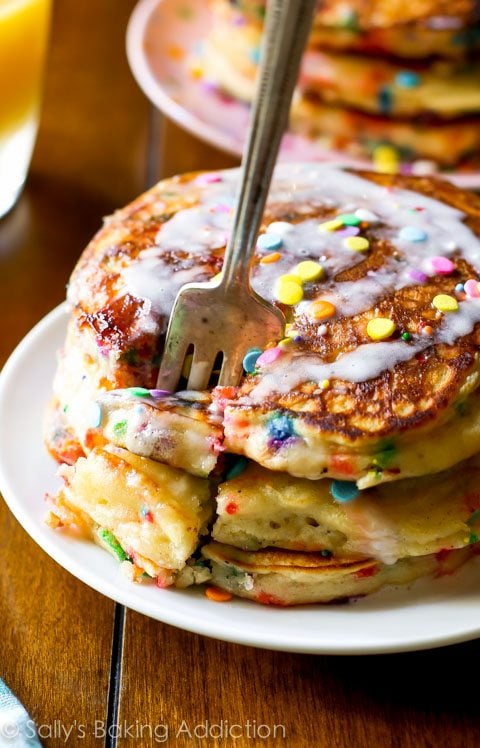 stack of funfetti buttermilk pancakes topped with vanilla icing and sprinkles on a white plate with a fork
