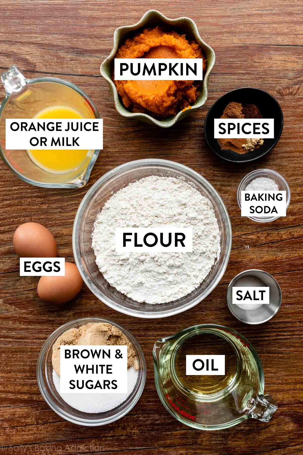 flour, baking soda, spices, salt, oil, eggs, and other ingredients in bowls on wooden backdrop.