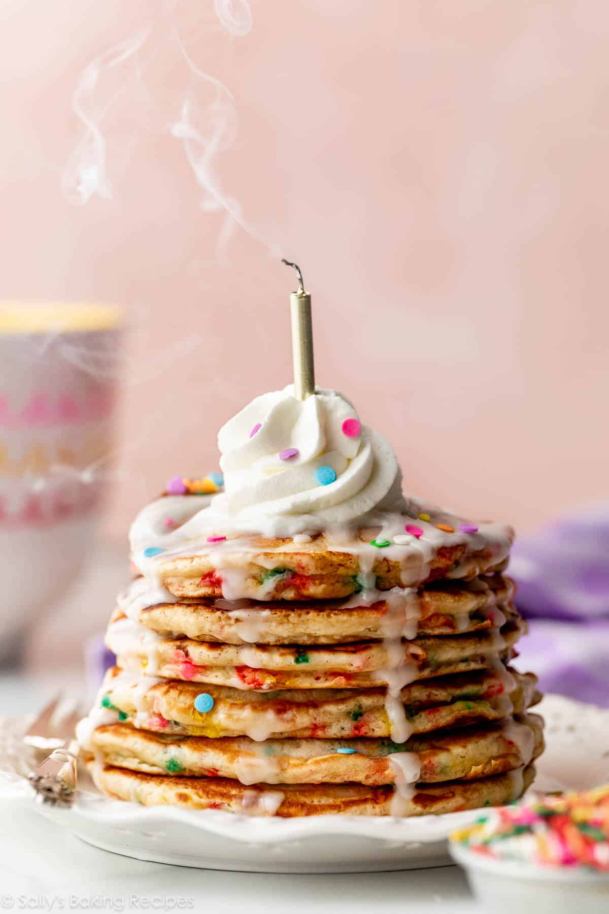 stack of birthday cake pancakes with icing dripping down the sides, whipped cream on top, and a candle on top that is smoking from just being blown out.