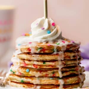 stack of birthday cake funfetti pancakes with icing dripping down the sides, whipped cream on top, and a lit gold candle on top.