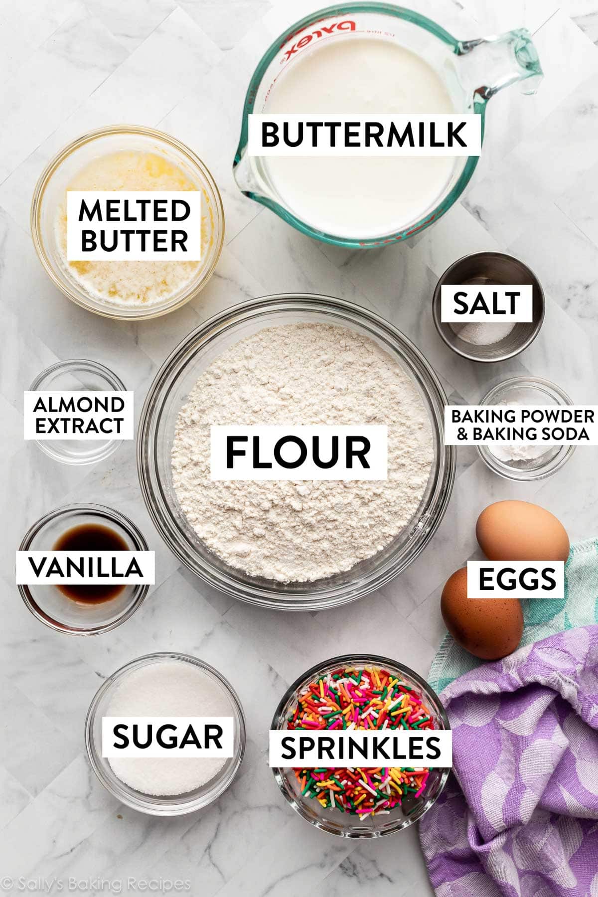 ingredients in bowls on marble counter including flour, sprinkles, salt, melted butter, vanilla, buttermilk, and sugar.
