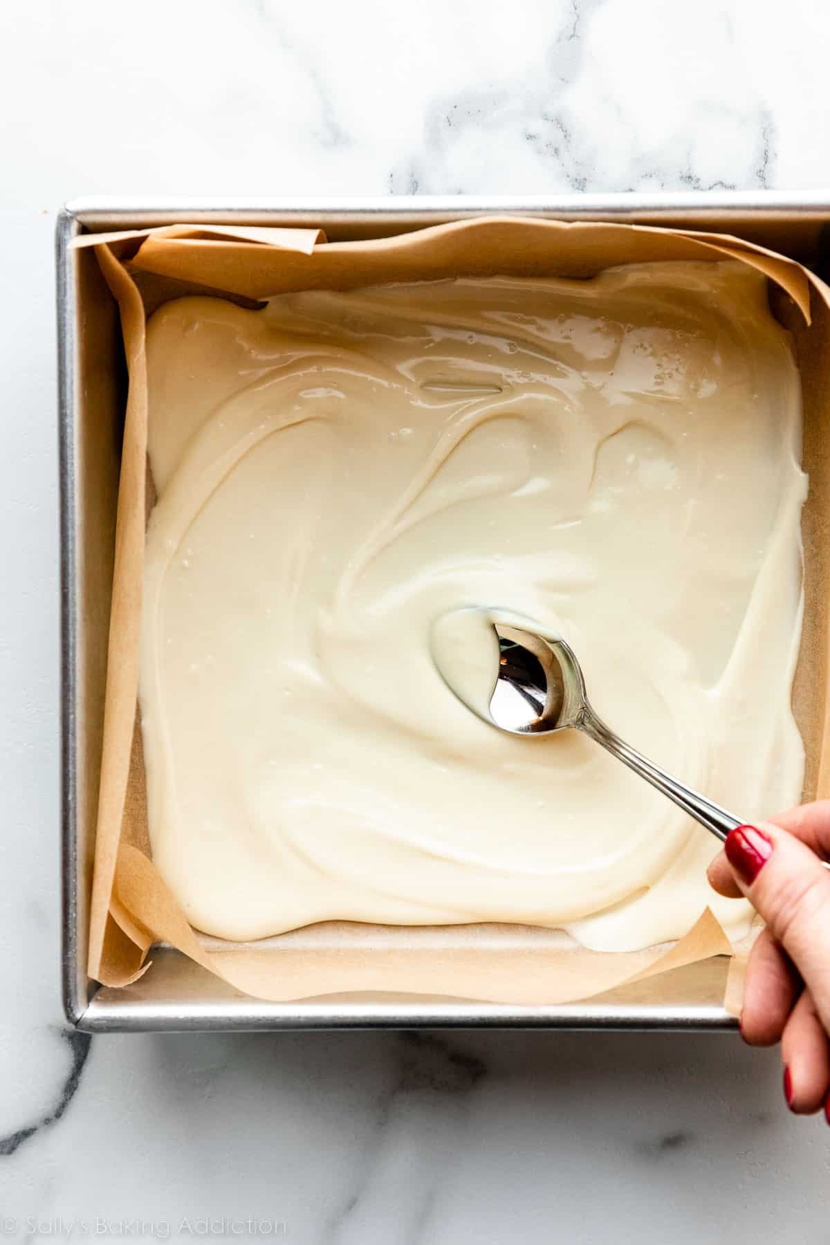spreading melted white chocolate with a spoon in lined square baking pan.
