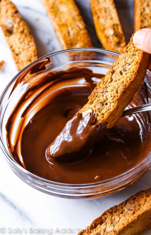 hand dipping almond biscotti into a glass bowl of melted chocolate