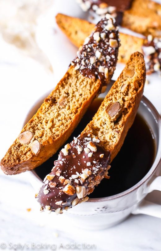 2 almond biscotti cookies with part of each cookie dipped in chocolate resting on top of a white cup of coffee