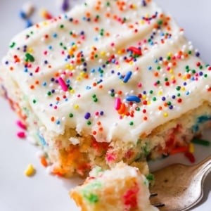 slice of funfetti sheet cake topped with vanilla frosting and sprinkles on a white plate with a fork