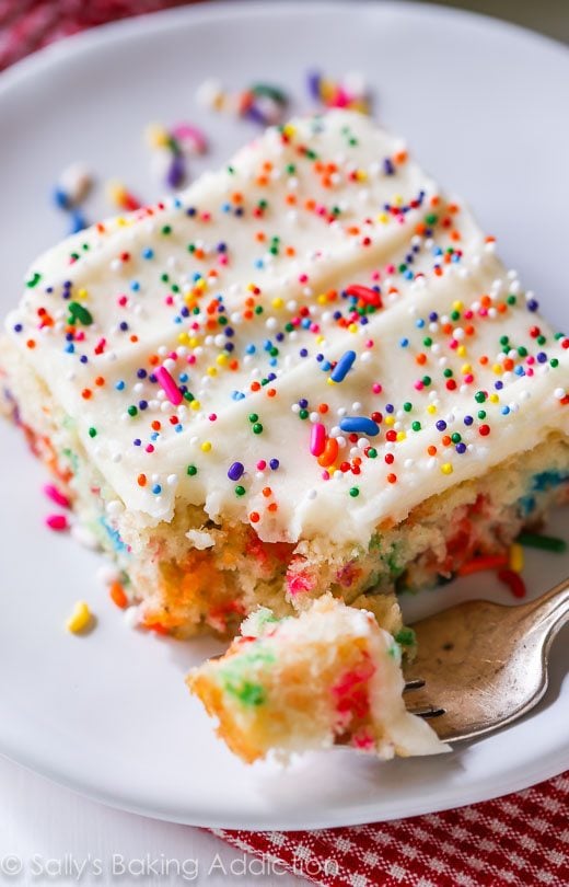 slice of funfetti sheet cake topped with vanilla frosting and sprinkles on a white plate with a fork
