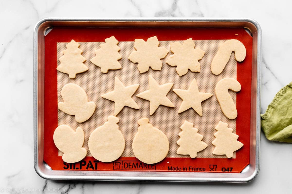 sugar cookies in various shapes on silicone mat-lined baking sheet including stars, candy canes, and snowflakes.