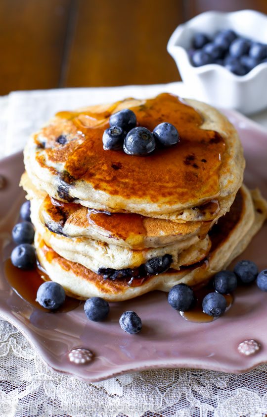 stack of whole wheat blueberry pancakes topped with maple syrup and fresh blueberries on a pink plate